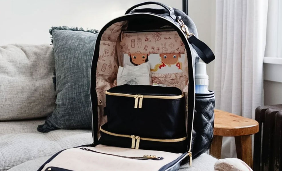the itzy ritzy boss diaper bag sitting on a couch with zipper open and diaper and baby book inside